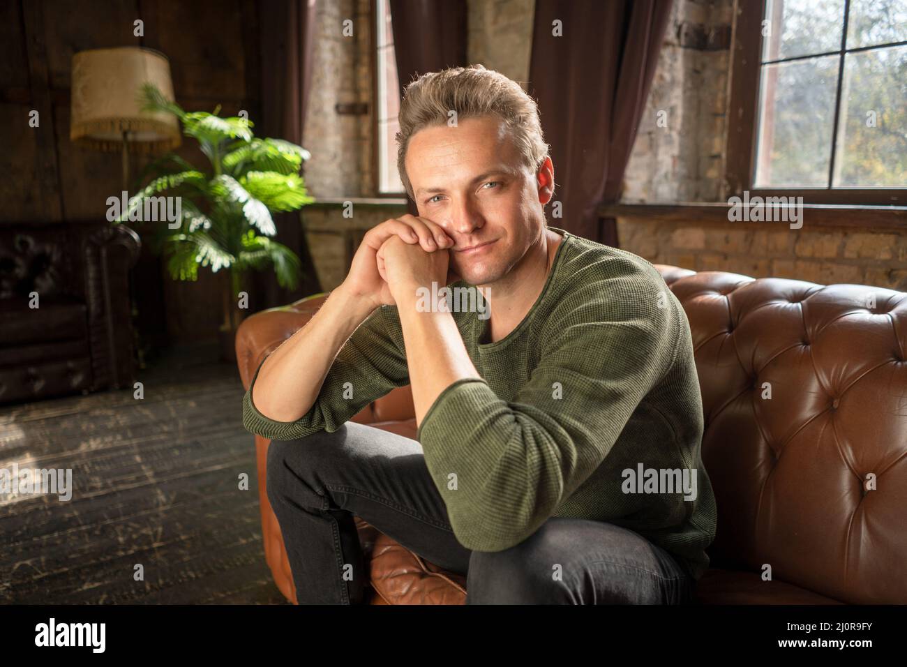 Attraktiver Mann von 35s Jahren mit liebevollen Augen in legerer Kleidung sitzt auf einem weichen Ledersofa in seinem luxuriösen Wohnzimmer seines Landes H Stockfoto