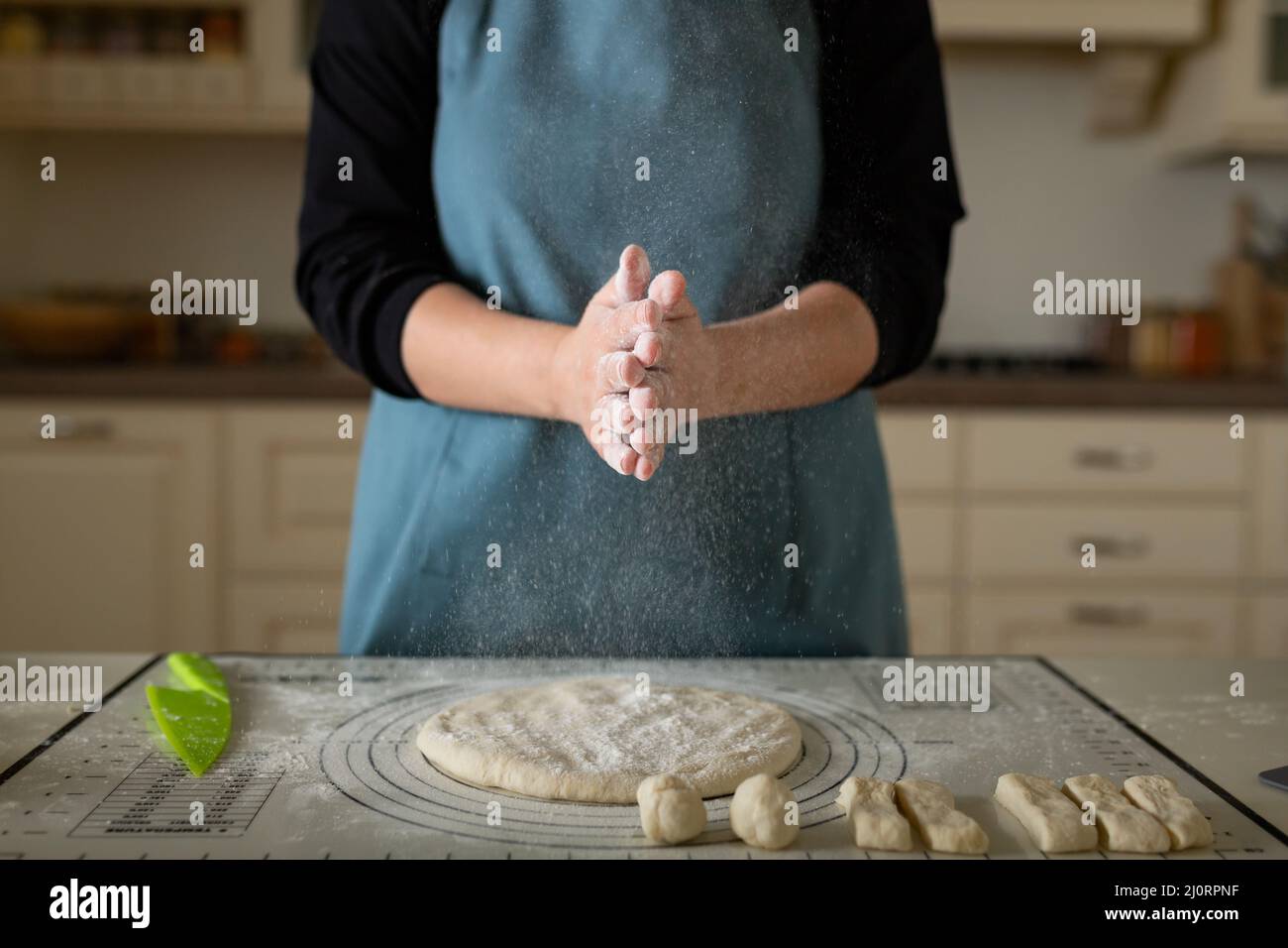 Klatschende Hände im Mehl eines Kochs in einer blauen Schürze in einer heimischen Küche. Stockfoto