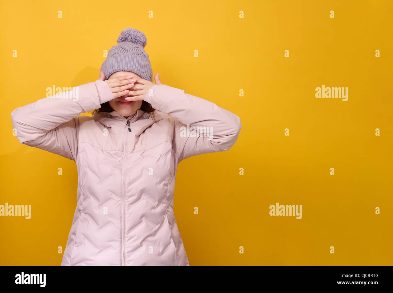 Eine 40-ige Frau, gekleidet in eine pinke Winterjacke und einen Hut, schließt ihre Augen mit ihren Händen auf einem gelben Hintergrund. Warten auf Stockfoto