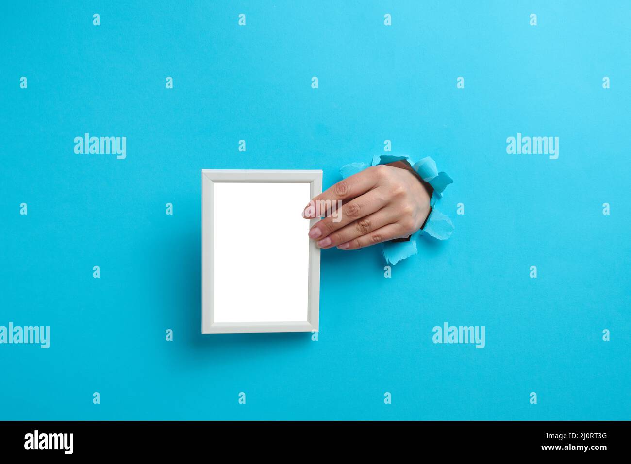 Weibliche Hand hält einen leeren weißen Holzrahmen, der Körperteil ragt aus dem zerrissenen Loch im blauen Papierhintergrund Stockfoto