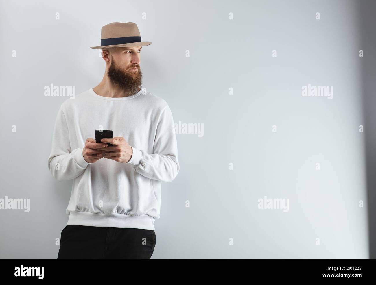 Ernsthafter bärtiger Mann im Hut, der mit einem Smartphone gegen eine weiße Wand steht. Stockfoto