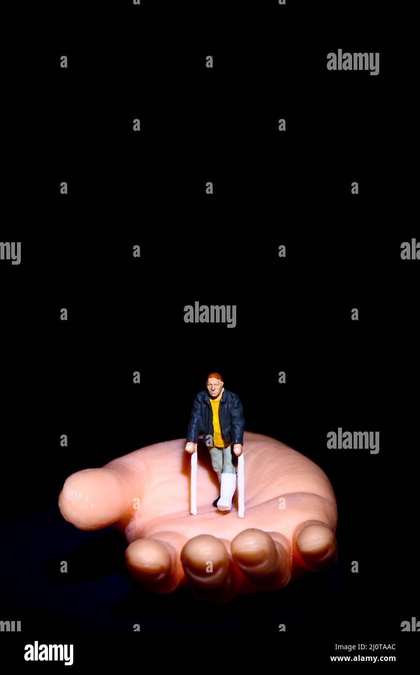 Miniaturfigur Mann mit Krücken saß auf einer gefälschten Hand deaktiviert helfende Hand Konzept mit Kopierraum Stockfoto