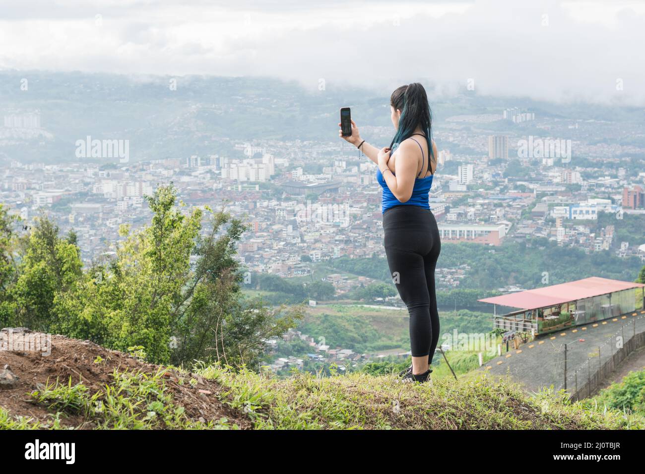 Junge Frau. lateinische Influencerin, die ein Video für ihre sozialen Netzwerke auf einem Berg aufzeichnet, im Hintergrund die Stadt Pereira-Kolumbien Stockfoto