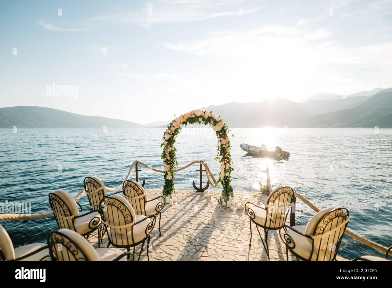 Reihe von weichen Stühlen vor einem Hochzeitsbogen auf einem gepflasterten Pier mit dem Meer und den Bergen im Hintergrund Stockfoto