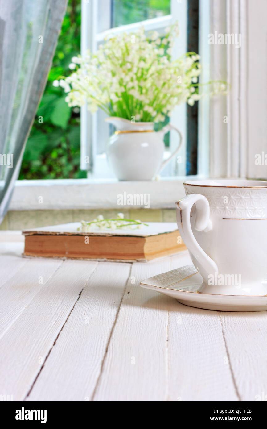 Eine Tasse Tee oder Kaffee und ein Buch auf einem weißen Retro-Holztisch und ein Blumenstrauß von Maiglöckchen auf dem Windo Stockfoto