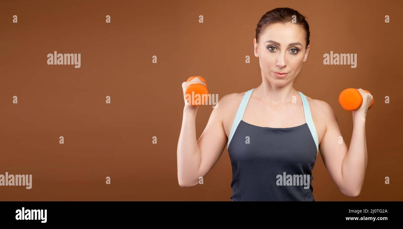 Panoramaaufnahme einer athletischen kaukasischen Frau mit Kurzhanteln in ihren Händen. Steht auf einem braunen Hintergrund mit Hanteln in seinem ha Stockfoto