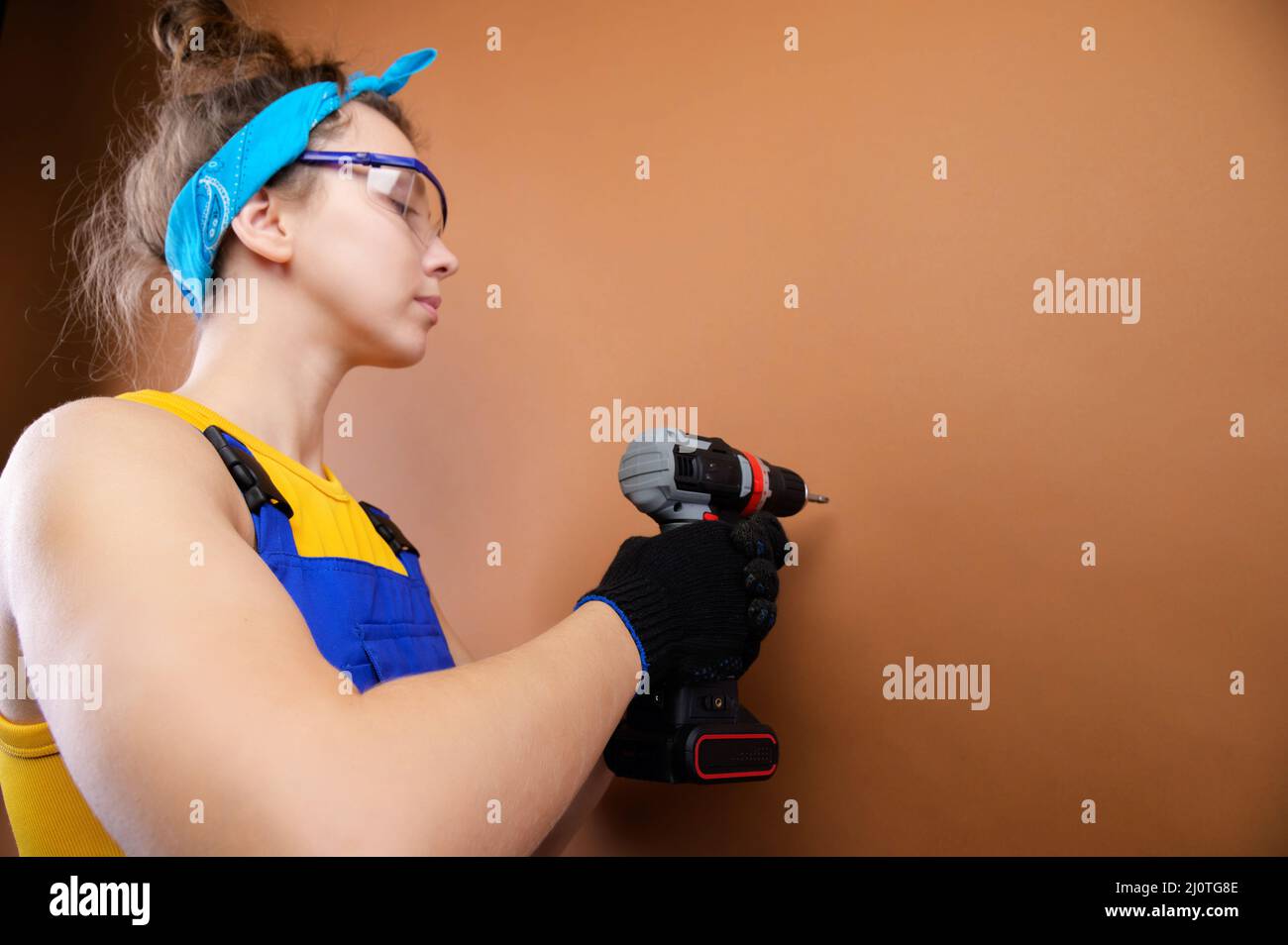 Junge Frau in Overalls und Brillen arbeitet mit einem Schraubendreher im Studio auf braunem Hintergrund. Schwerpunkt auf die Schraube legen Stockfoto