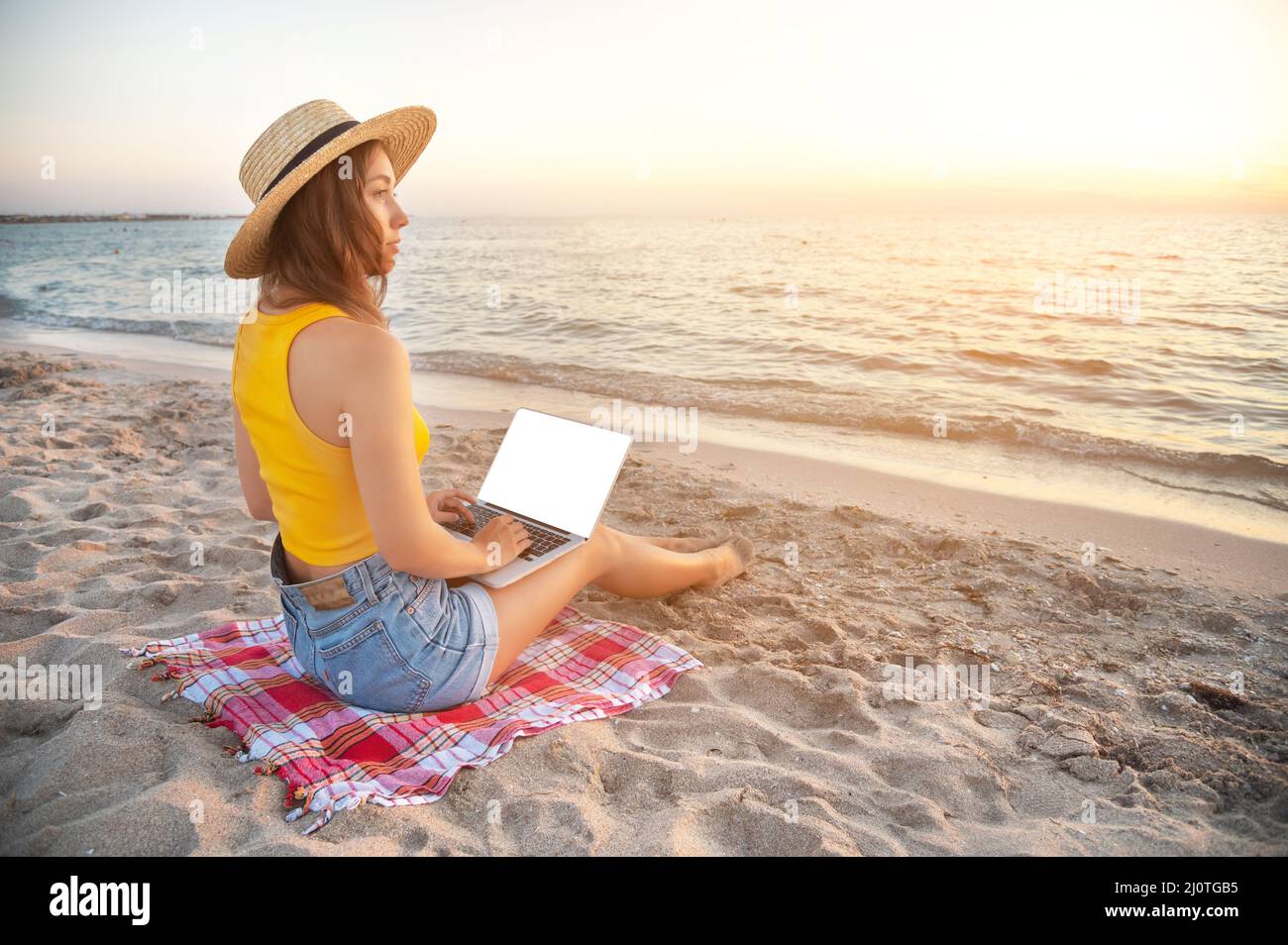 Junge kaukasische Frau in Shorts und einem Hut am Strand, sitzt auf dem Sand, browst soziale Netzwerke auf dem Computer im Sommer ein Stockfoto