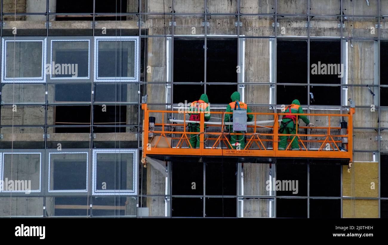 Die Arbeiter in der Bauwiege in der Höhe sind im Winter mit der Verglasung der Fassade eines Wolkenkratzers beschäftigt. Stockfoto