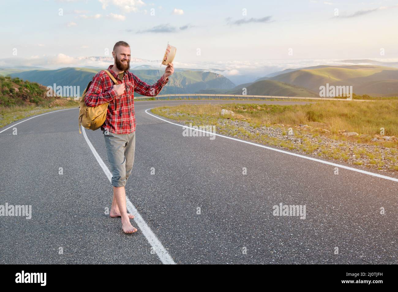 Ein lächelnder, bärtiger, freundlicher kaukasischer Mann in einem Hemd und Shorts mit Rucksack begrüßt Sie, indem er seinen Hut hebt, während er auf einem c steht Stockfoto