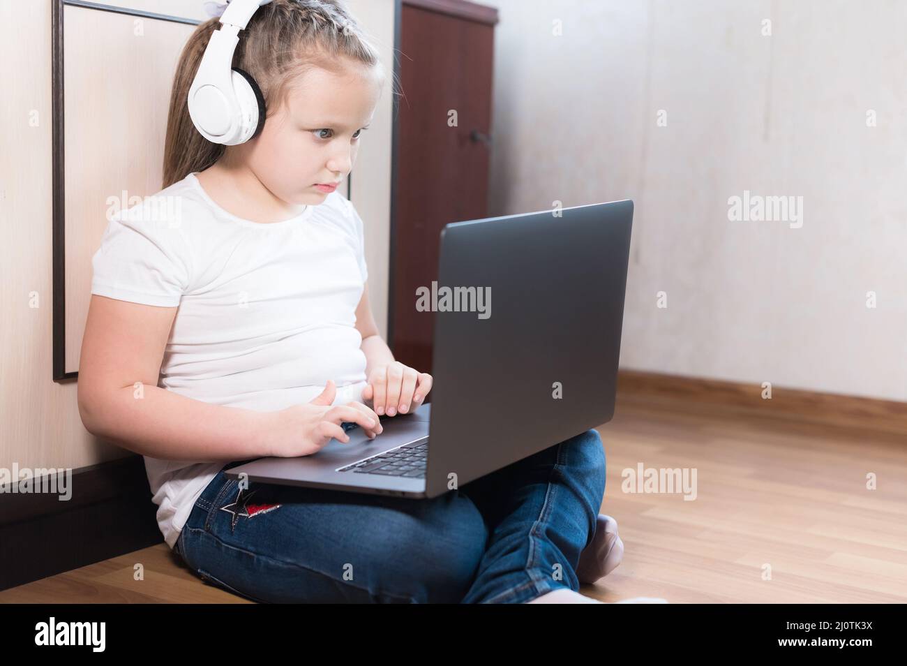 Ein junges kaukasisches Mädchen in einem weißen T-Shirt, Kopfhörern und Jeans sitzt auf dem Boden in einem Raum mit einem Laptop in den Armen. Ein Stockfoto