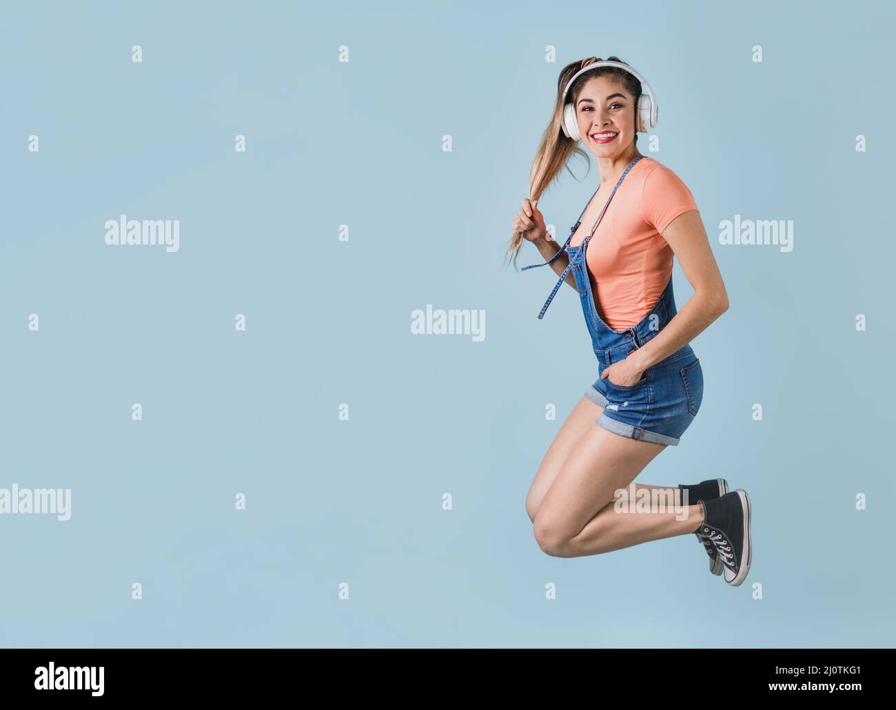 hispanische junge Frau mit Kopfhörern, die springen und Musik auf blauem Hintergrund hören in Mexiko, Lateinamerika Stockfoto