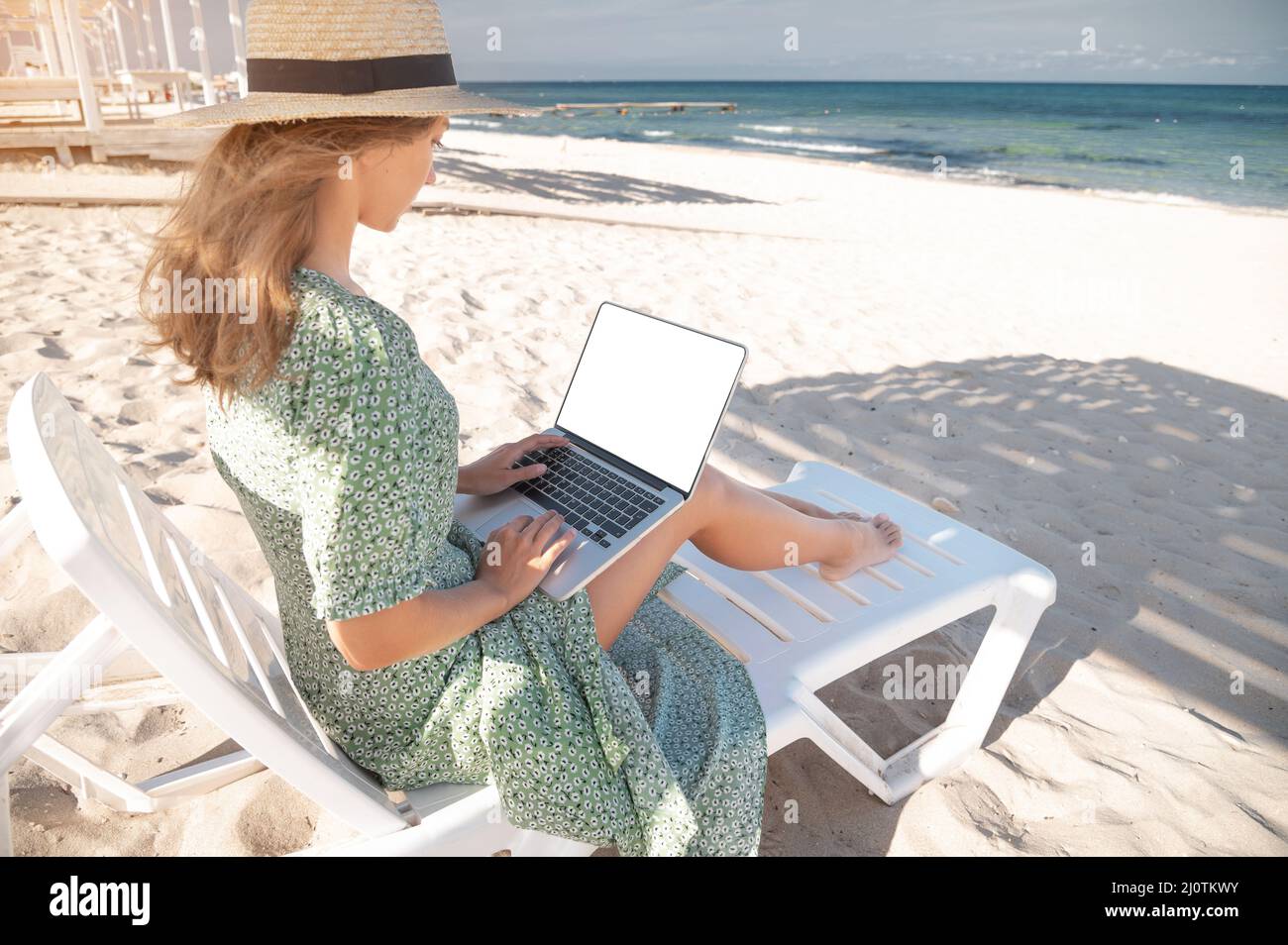Rückansicht einer attraktiven jungen kaukasischen Frau in einem grünen Kleid und Hut sitzt auf einer Liege mit einem Laptop in den Händen in der sh Stockfoto