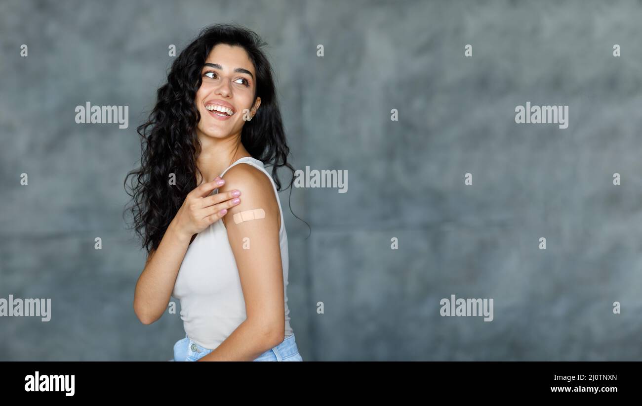 Coronavirus-Impfung. Glückliche junge Frau, die Bandhilfe auf ihrem Arm zeigt und auf grauen Hintergrund auf freien Raum schaut Stockfoto