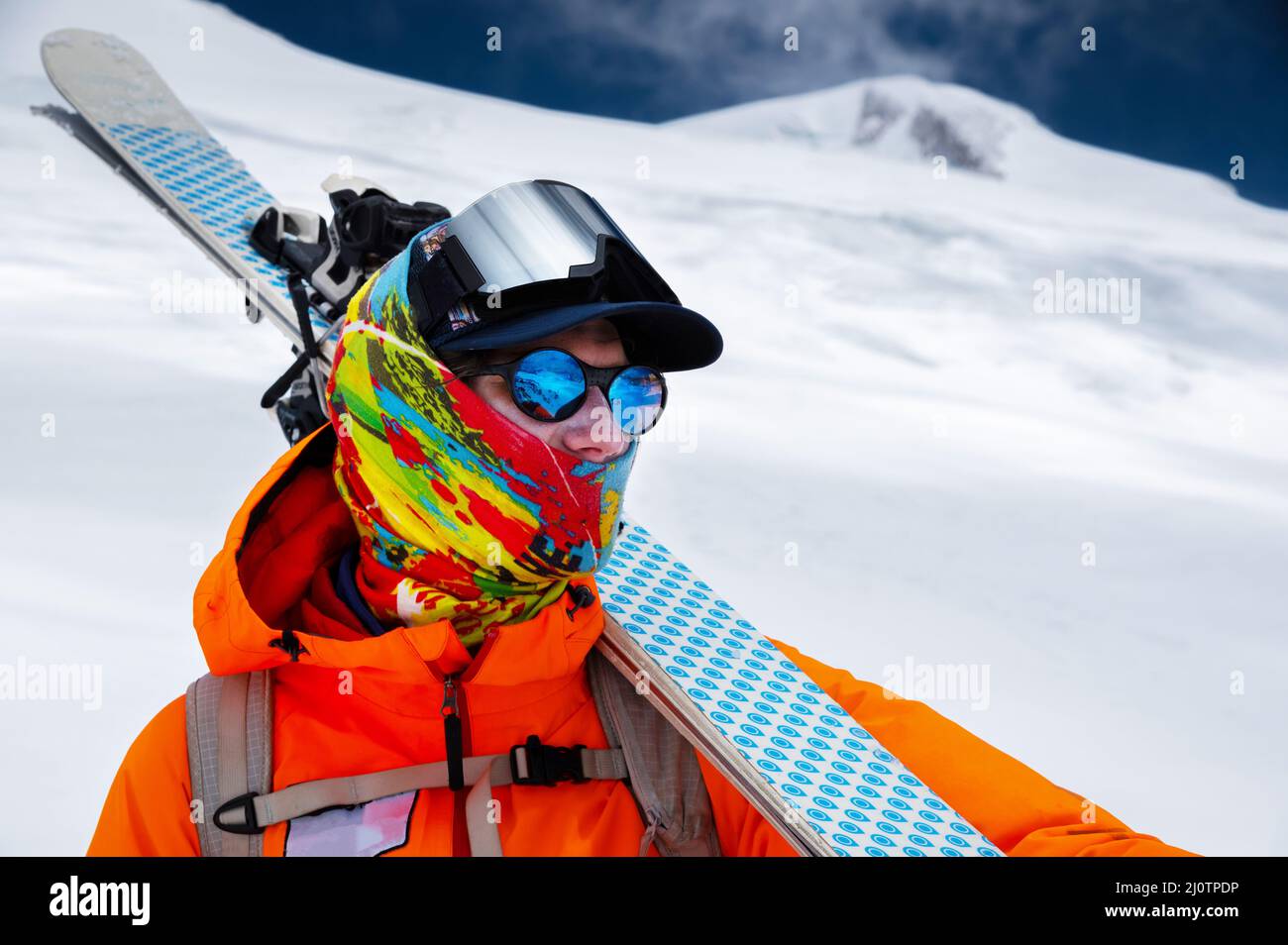 Bergporträt eines professionellen Freeridskiers in orangefarbener Kleidung mit Skistöcken und Skiern auf den Schultern. Steht hoch in t Stockfoto