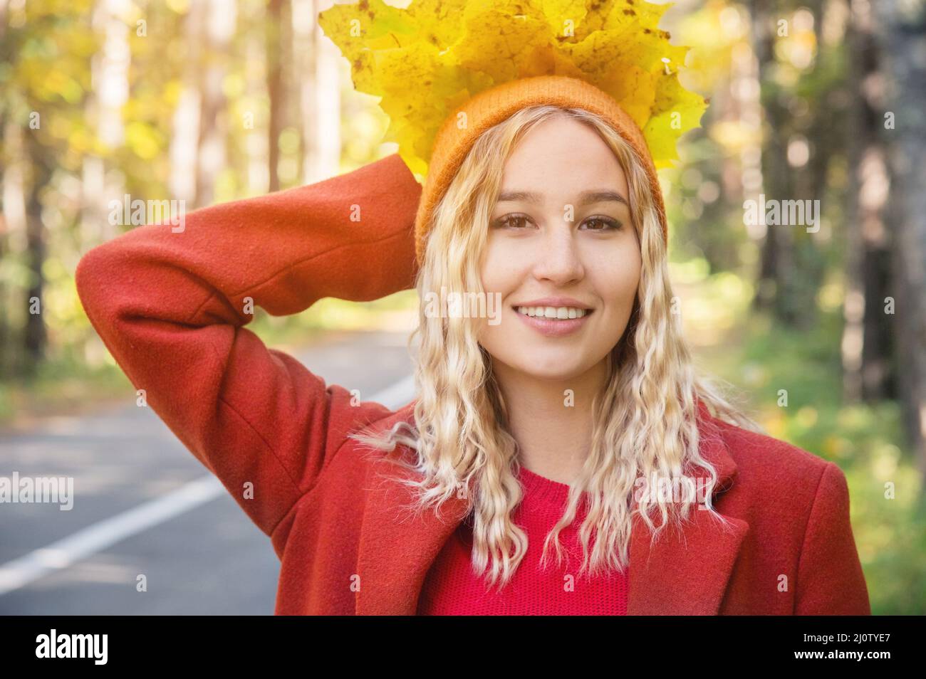 Attraktives blondes Mädchen mit lockigem Haar in orangefarbener Mütze und rotem Mantel hält einen Strauß gelber Blätter und lächelt gegen die Roa Stockfoto