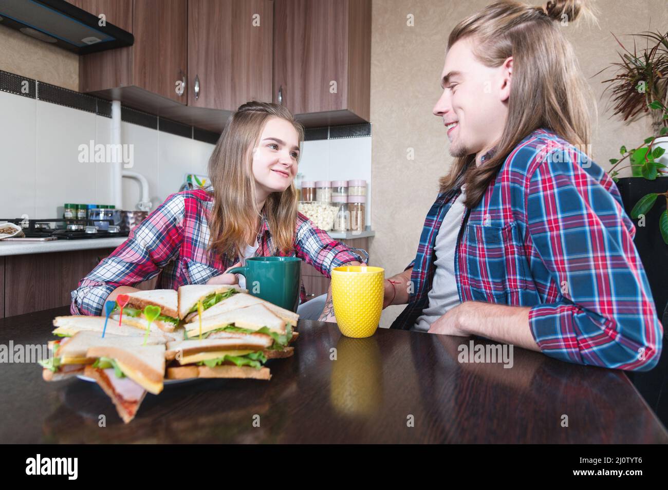 Eine junge Familie trinkt Tee mit Sandwiches in der Küche am Esstisch und schaut sich gegenseitig an. Ein Ehepaar mit langen ha Stockfoto