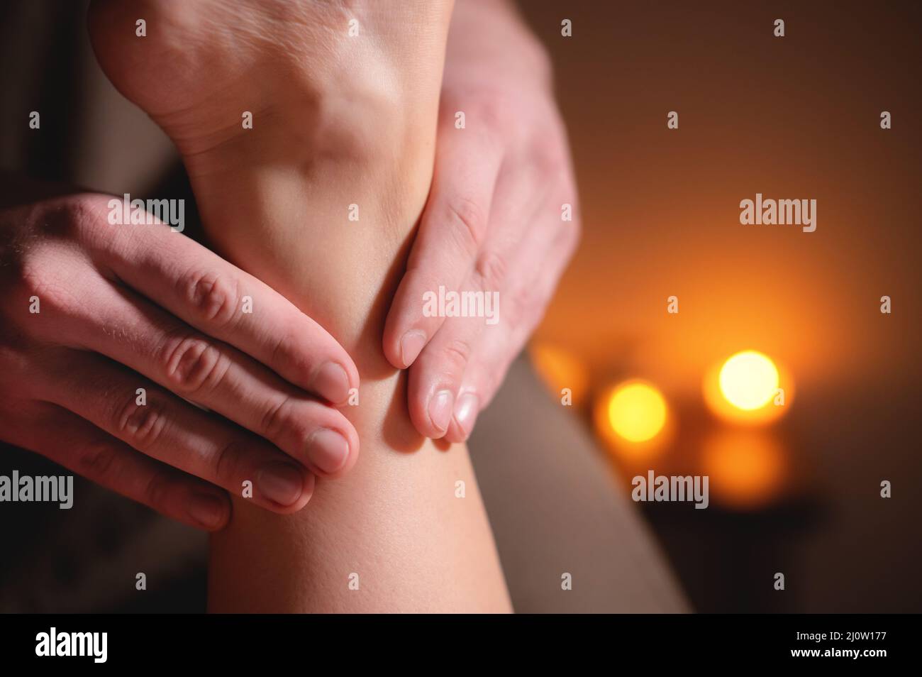 Nahaufnahme eines männlichen Masseurs, der in einem dunklen Raum eine Bein- und Fußmassage vor dem Hintergrund einer professionellen Kerzenmassage macht Stockfoto