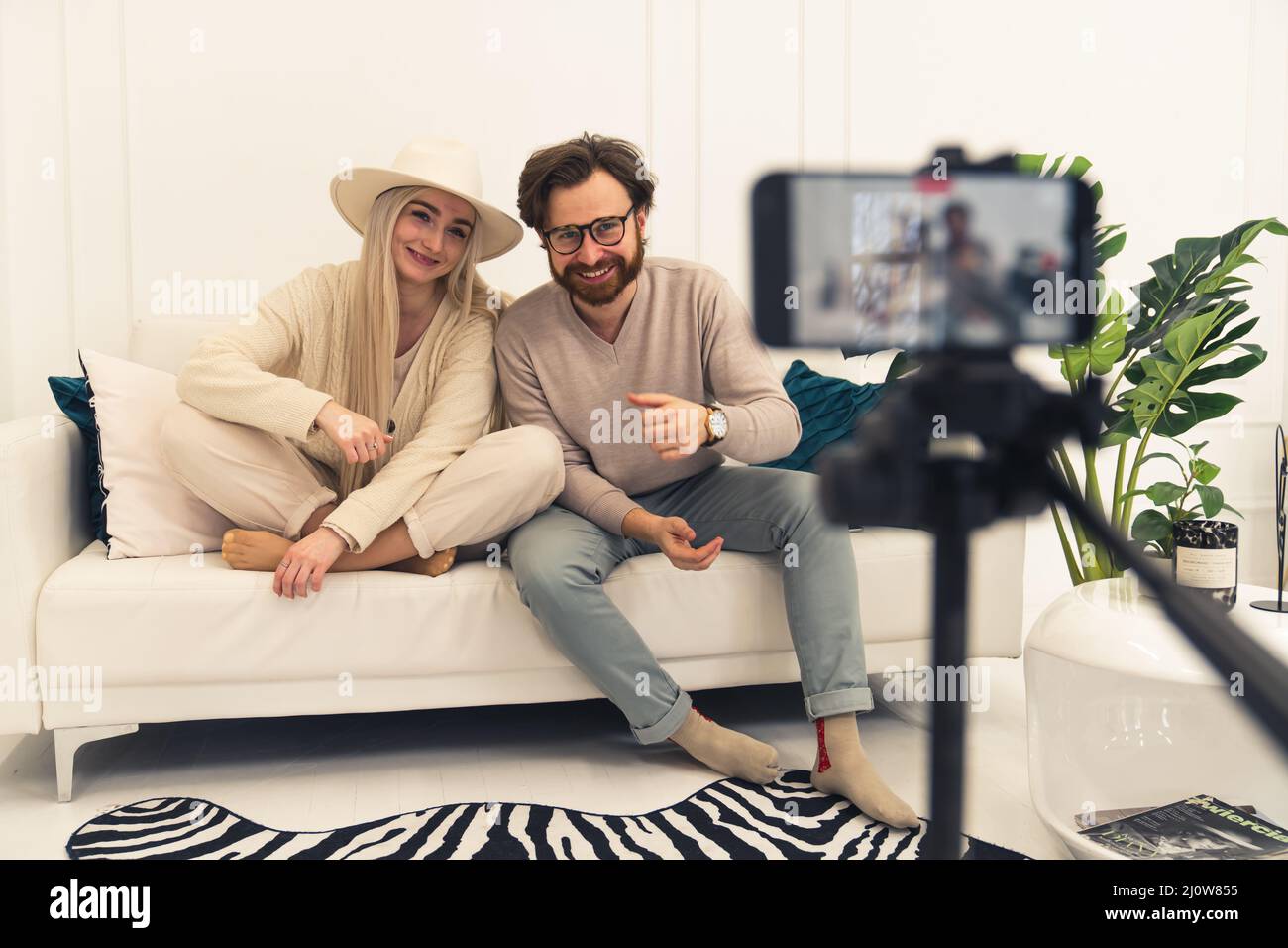 Junge verheiratete lächelndes kaukasisches Paar, das Videoinhalte mit einem Smartphone auf der Couch im Innenbereich macht. Hochwertige Fotos Stockfoto
