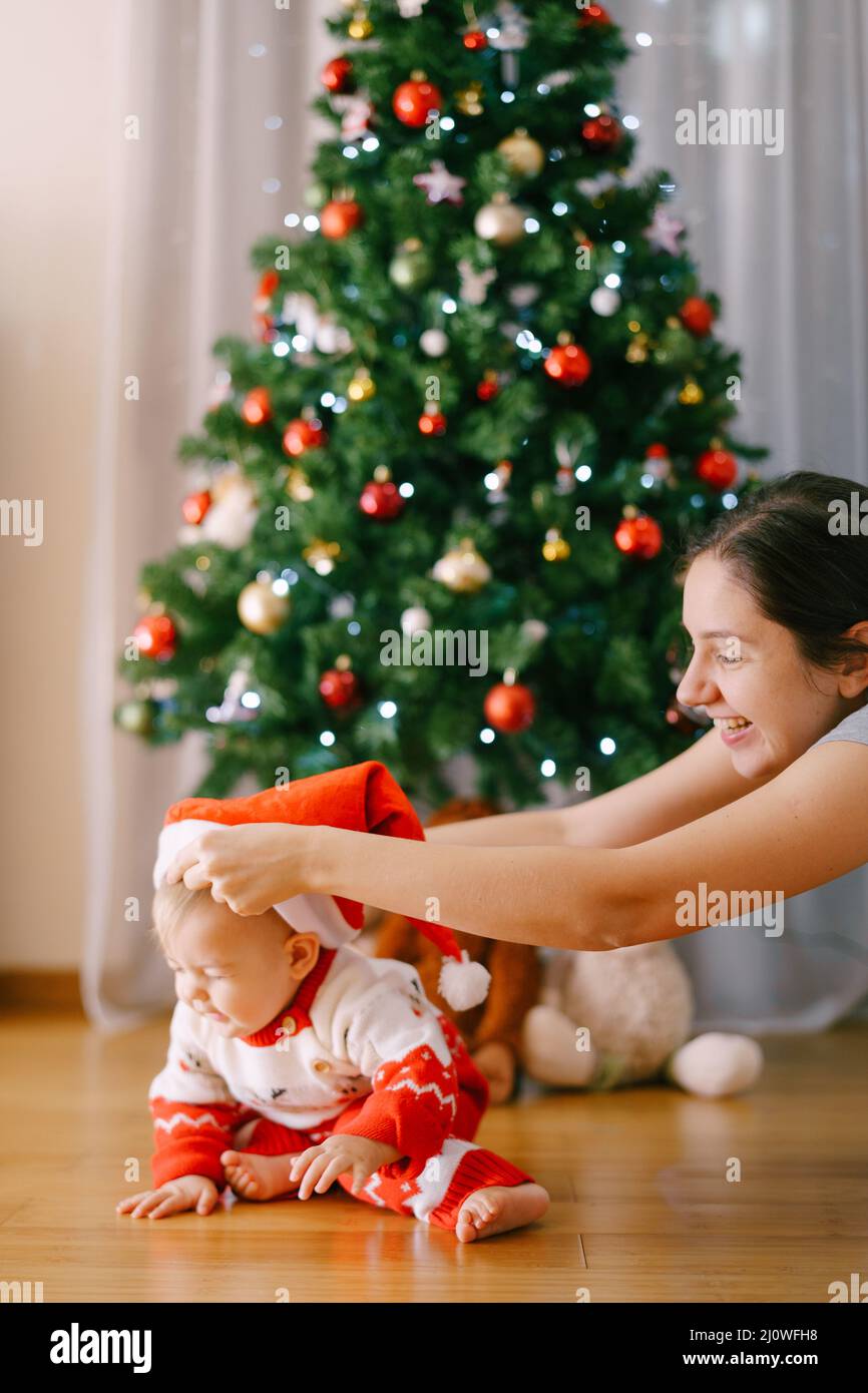Mutter setzt Weihnachtsmütze auf ihre kleine Tochter Vor einem Weihnachtsbaum Stockfoto