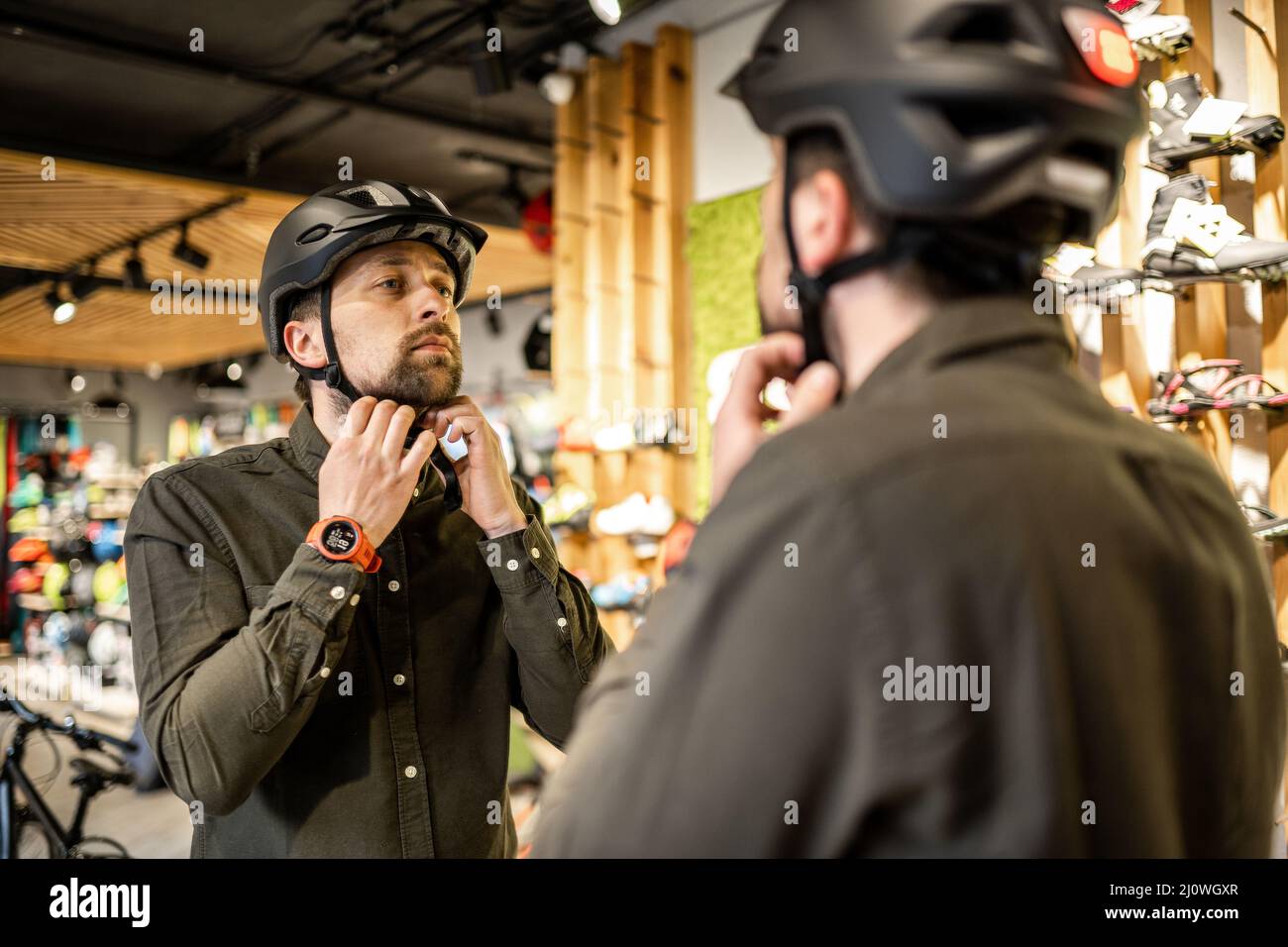 Mann, der Fahrradhelme im Sportgeschäft untersucht und sich im Spiegel ansieht. Neue Sporthelme im Fahrradladen ausprobieren. Mann setzt sich an Stockfoto