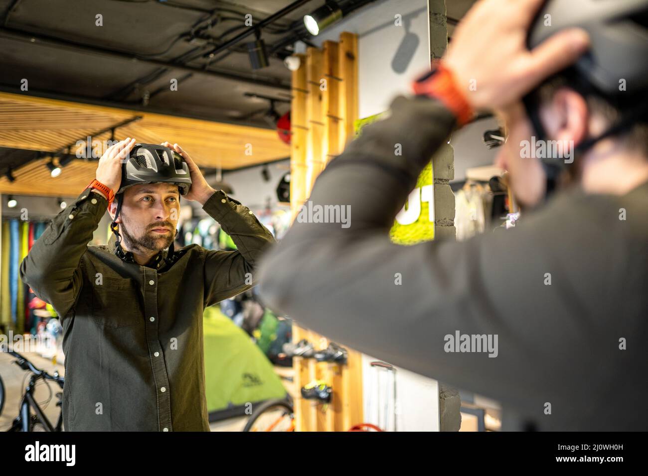 Kaukasischer Mann versucht auf schwarzem Fahrradhelm in der Nähe des Spiegels in Sportartikelgeschäft. Männlicher Käufer wählt Sicherheitshelm für Radfahren. Stockfoto