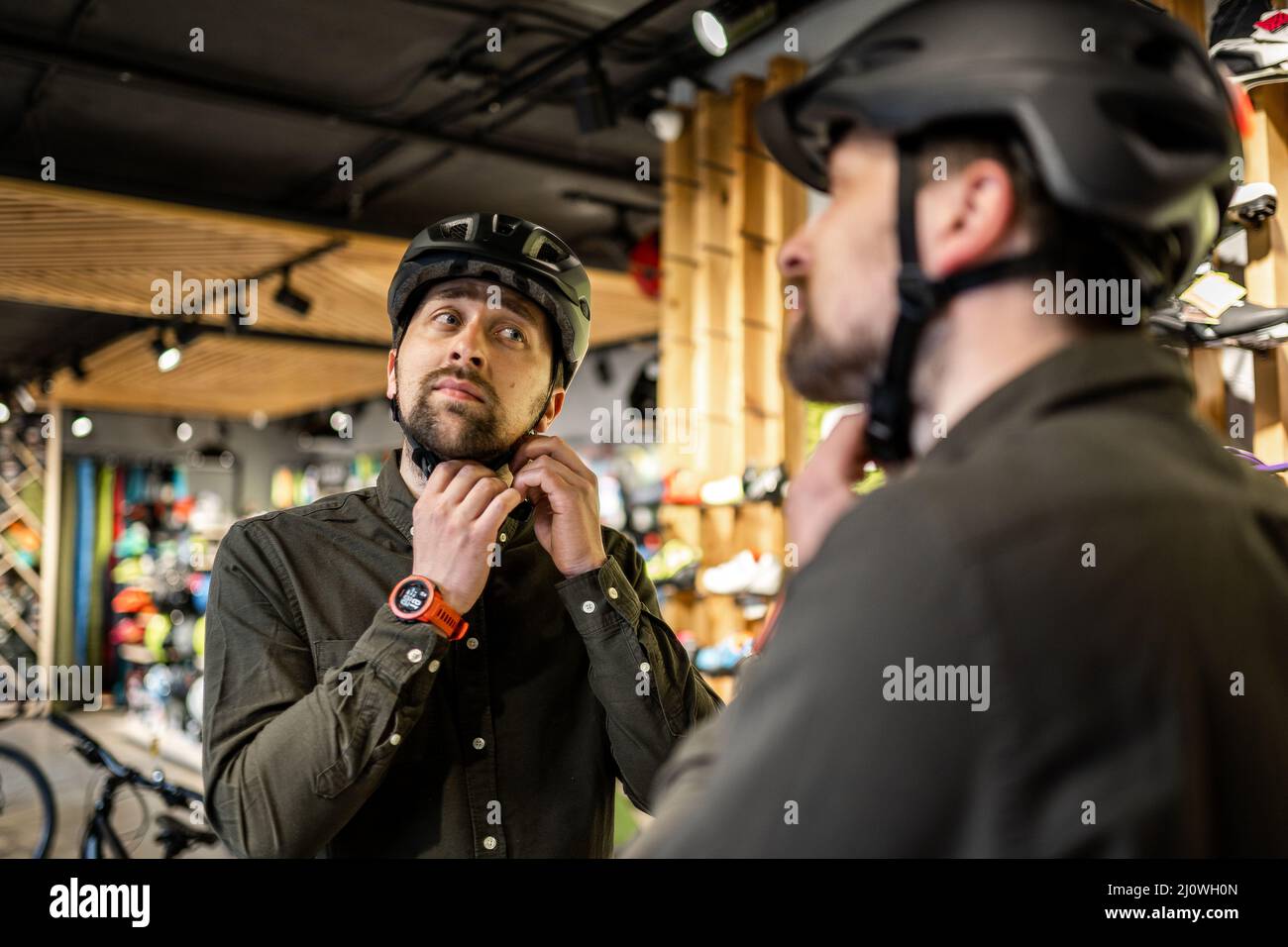 Kaukasischer Mann versucht auf schwarzem Fahrradhelm in der Nähe des Spiegels in Sportartikelgeschäft. Männlicher Käufer wählt Sicherheitshelm für Radfahren. Stockfoto