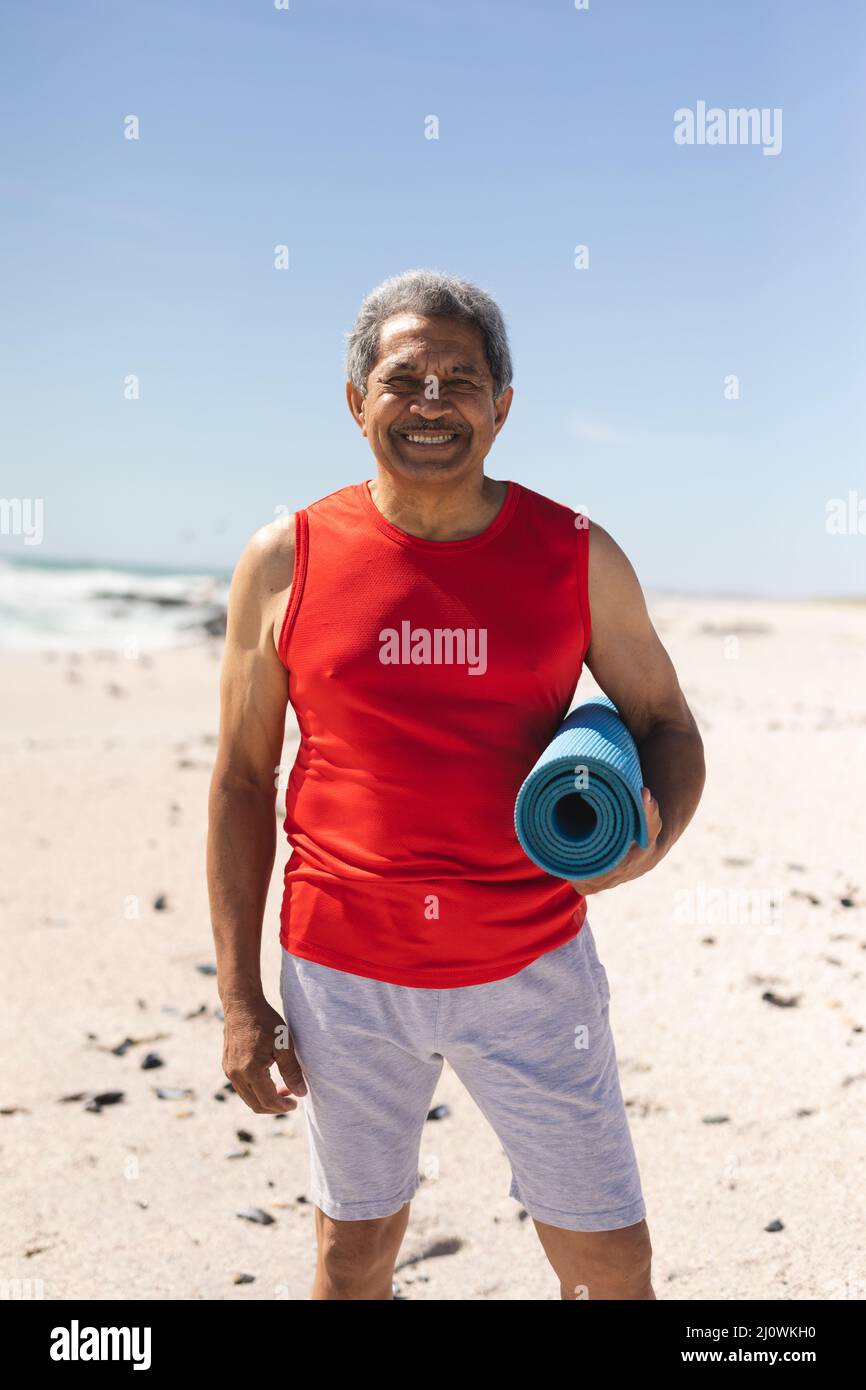 Porträt eines lächelnden pensionierten Birazialmannes, der am Strand eine gerollte Yogamatte gegen den blauen Himmel hält Stockfoto