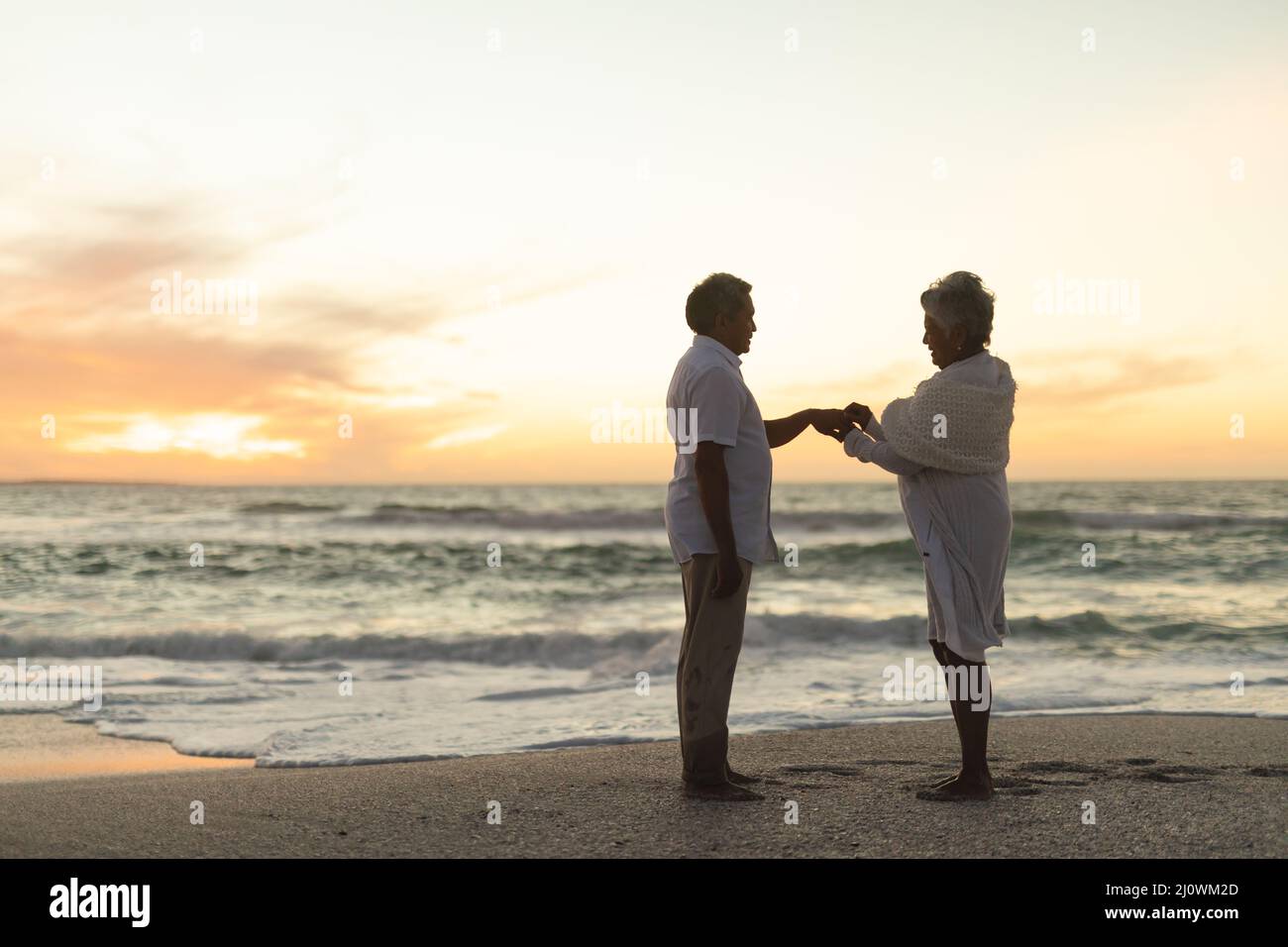 Seitenansicht der älteren biracial Braut, die Ehering auf den Finger des Bräutigams am Strand während des Sonnenuntergangs setzt Stockfoto