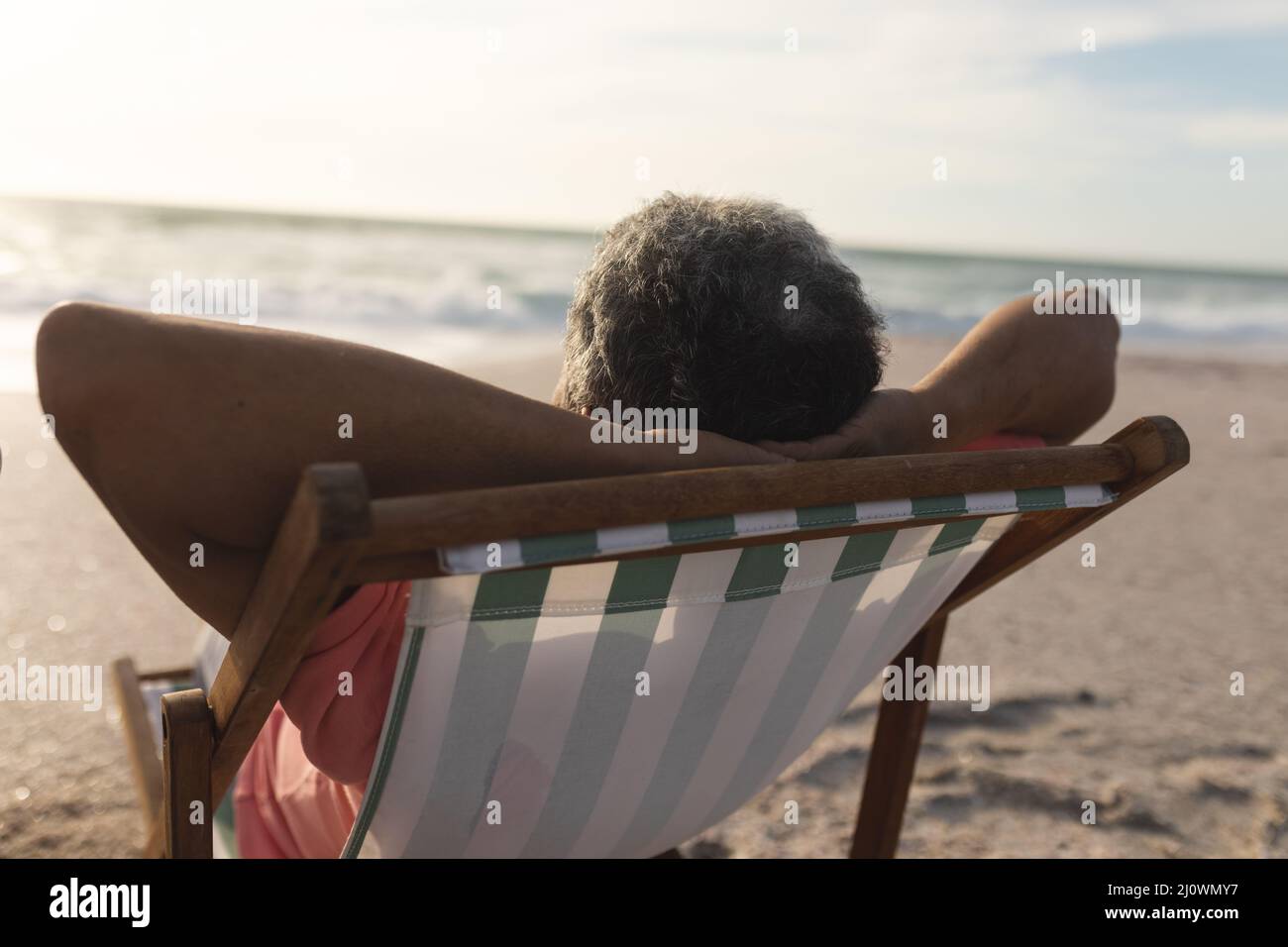 Der ältere Biracial-Mann entspannt sich während des Sonnenuntergangs mit den Händen hinter dem Kopf auf einem Klappstuhl am Strand Stockfoto