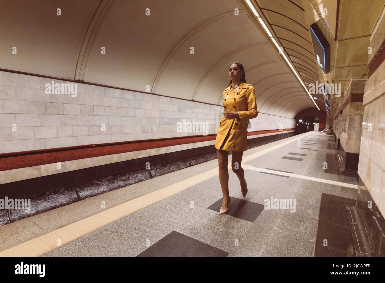 Auf der U-Bahn-Plattform spazieren schönes junges Mädchen mit langen Beinen in einem gelben Frühlingsmantel und einer weißen Handtasche in der Hand. Stockfoto