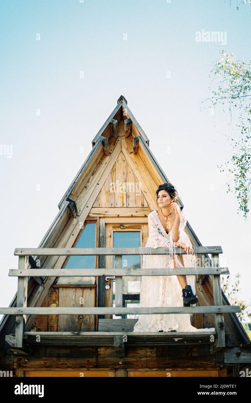 Die Braut in einem weißen Kleid steht auf dem Balkon eines dreieckigen Hauses und lehnt sich an ein Holzgeländer Stockfoto