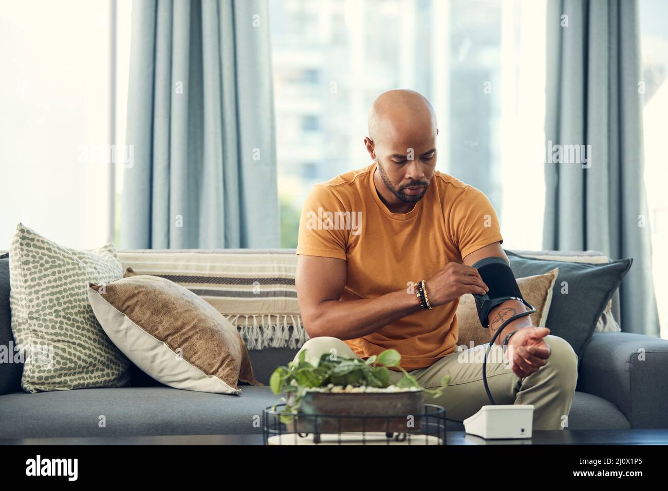 Ich habe Kopfschmerzen, so dass mein Blutdruck hoch sein könnte. Aufnahme eines jungen Mannes, der seinen Blutdruck nahm, während er zu Hause auf dem Sofa saß. Stockfoto