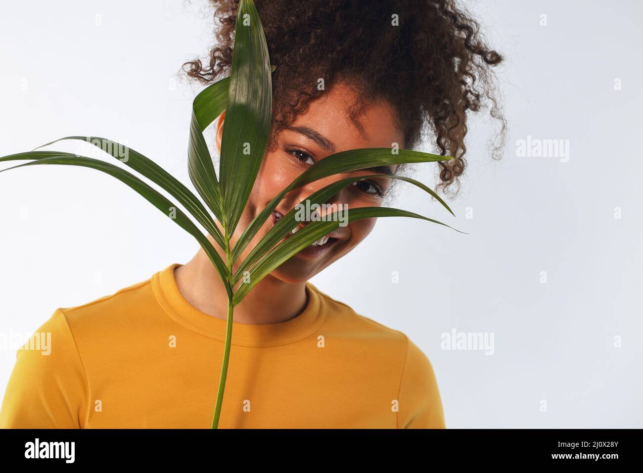 Junge fröhliche afroamerikanische Frau mit hoher Frisur, die tropische Pflanze im Gesicht hält und lächelt. Stockfoto