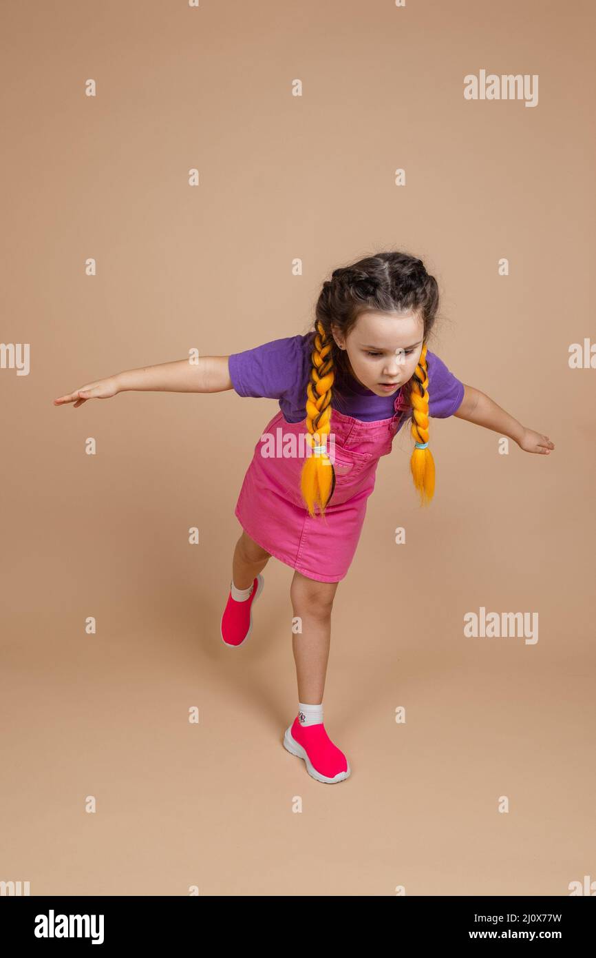 Portrait von verspielten kleinen Mädchen, mit gelben Kanekalon Zöpfe stehen auf einem Bein mit den Händen an den Seiten fliegen mit Augen nach unten tragen rosa gesetzt Stockfoto