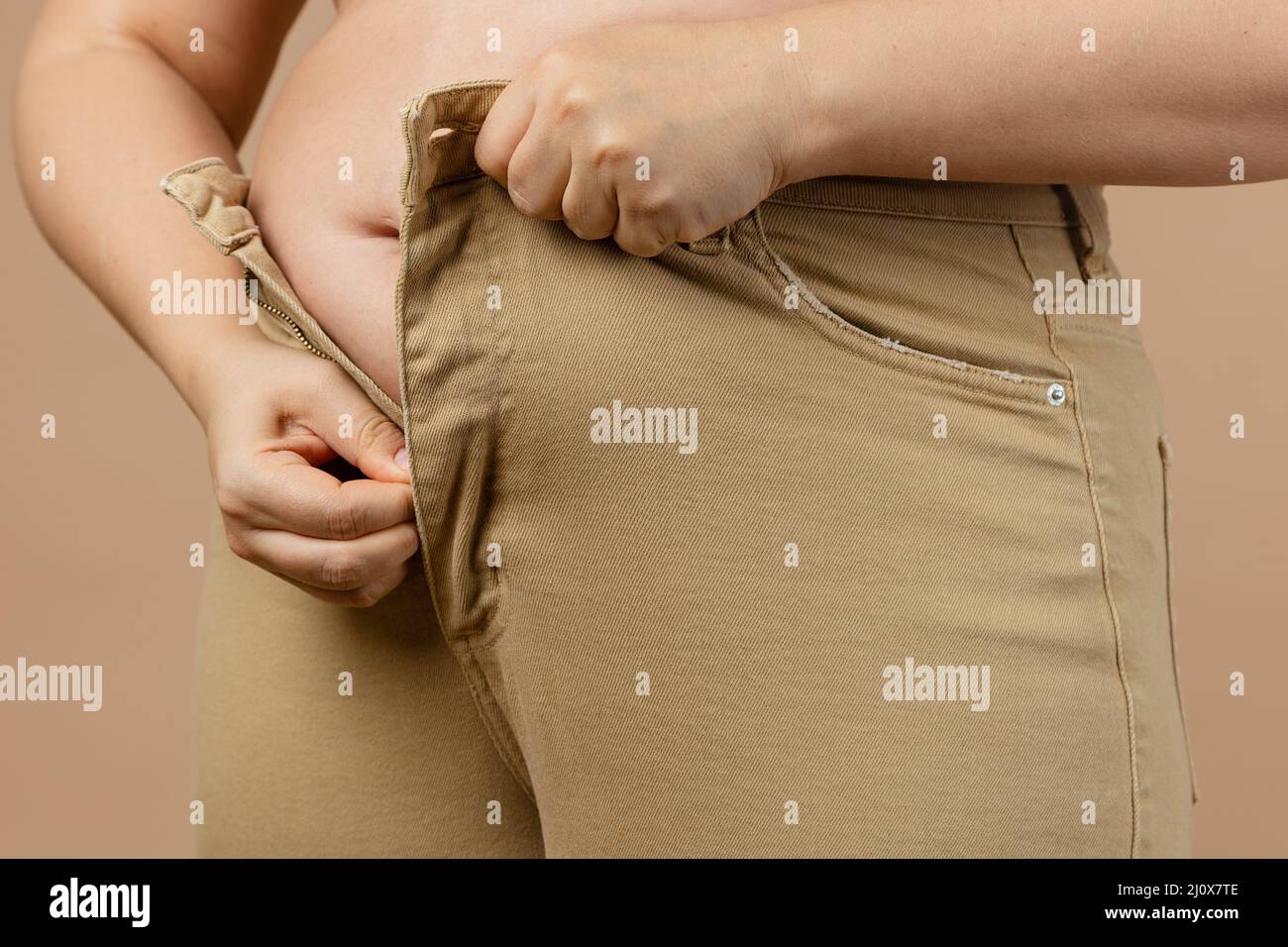 Frau großen Bauch nicht erlaubt ihr, Jeans auf beigem Hintergrund zu tragen. Viszerales Fett. Körper positiv. Plötzliche Gewichtszunahme. Enge Klamotten. Bedarf Stockfoto