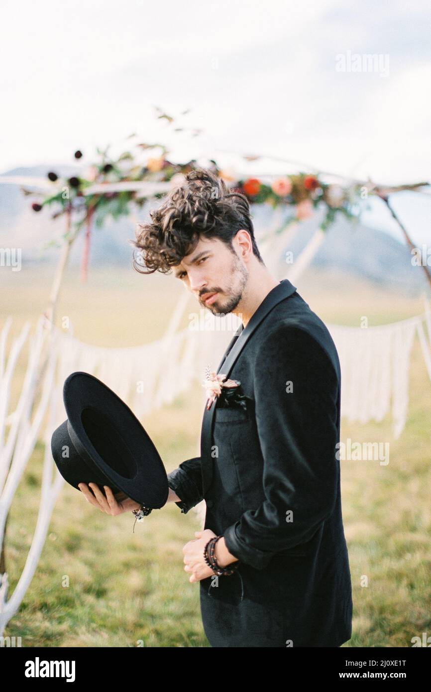 Der Bräutigam steht in einer schwarzen Jacke mit einem Hut in der Hand neben dem Hochzeitsbogen auf dem Feld Stockfoto