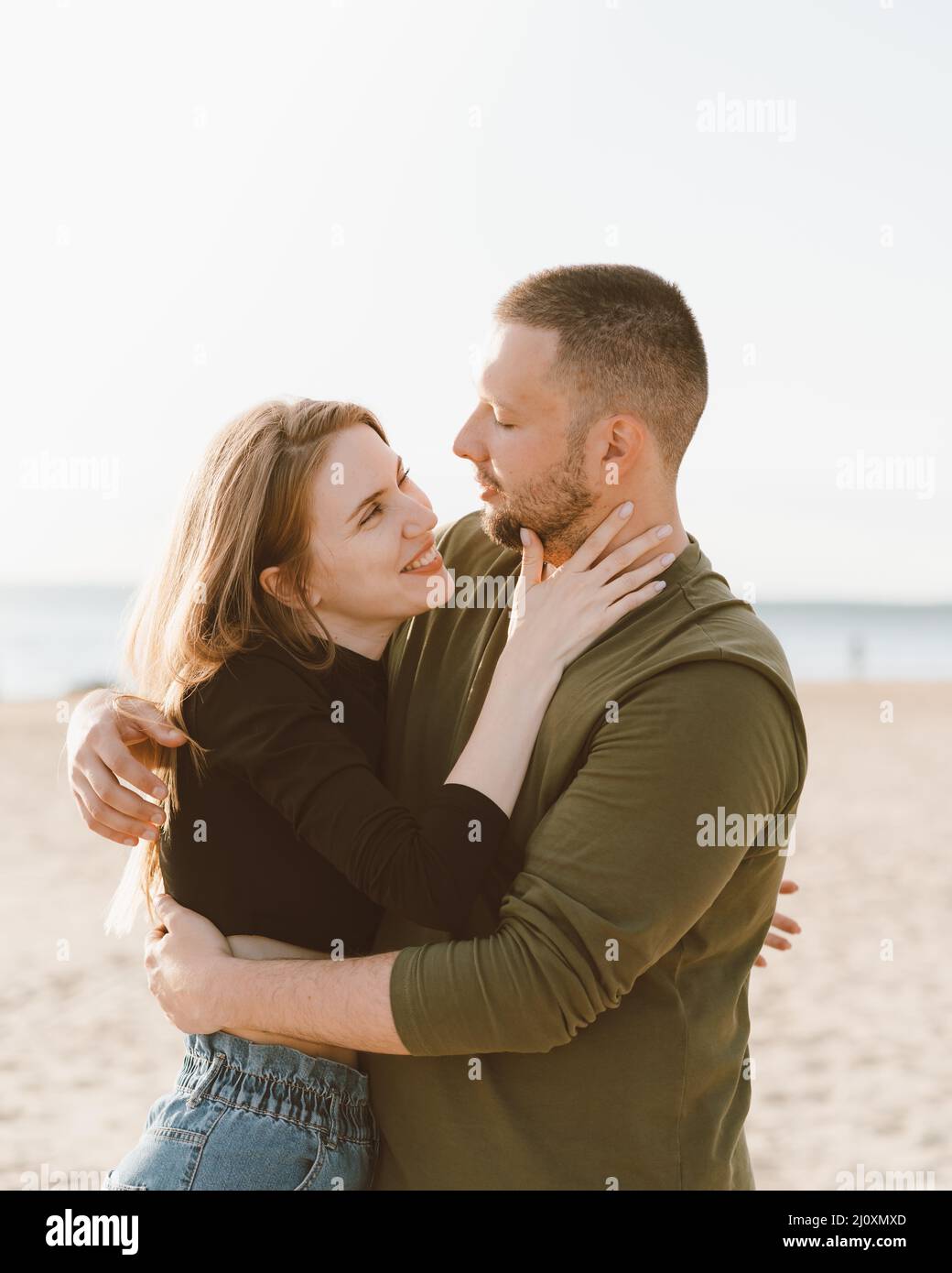 Junges erwachsenes Paar, das am Strand steht und sich gegenseitig schaut. Mann umarmt lachende Frau Stockfoto
