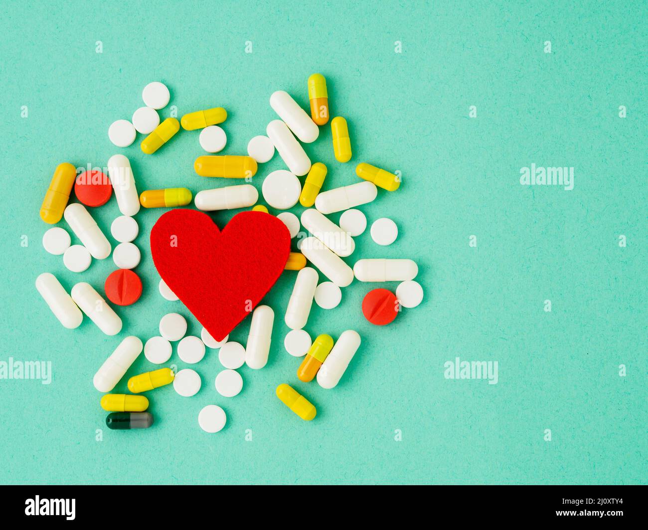 Eine Handvoll Pillen und ein rotes Herz auf einem leuchtend türkisblauen Farbtisch, Draufsicht, Kopierraum. Medizin hilft für die Gesundheit des Herzens. Stockfoto