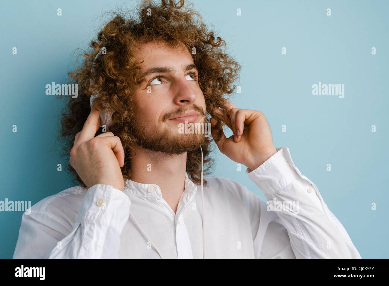 Junger lockiger Mann mit Ingwerhaar lächelt, während er Kopfhörer auf blauem Hintergrund verwendet Stockfoto