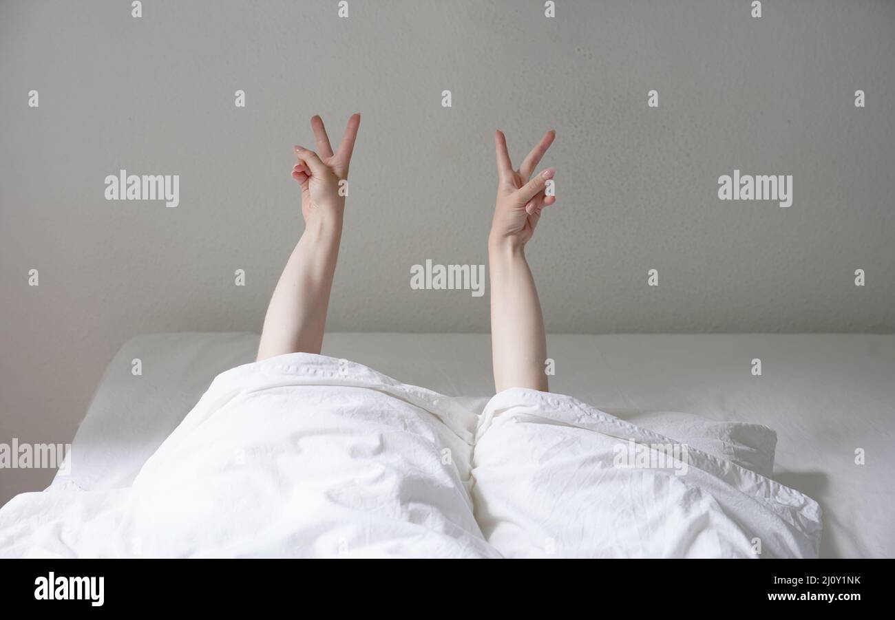 Frau, die in einem gemütlichen Bett liegt und sich unter den Decken versteckt, mit einem Friedenszeichen oder einem V-Handzeichen Stockfoto
