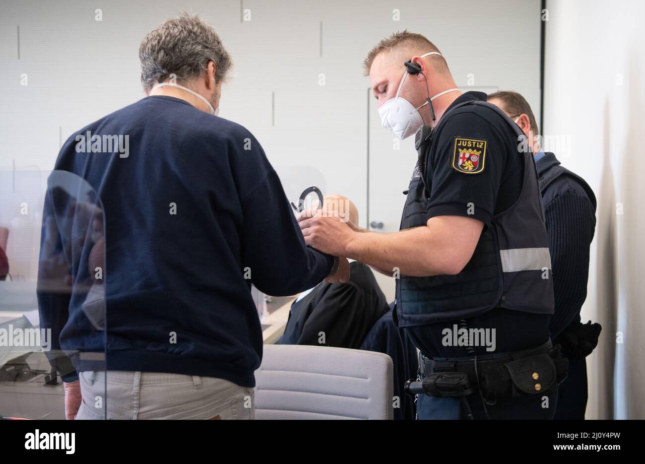 Bad Kreuznach, Deutschland. 21. März 2022. Der Angeklagte wird in Handschellen in den Gerichtssaal gebracht. Ein Justizvollzugsbeamer nimmt ihm die Handschellen ab. Der Mann soll im vergangenen Jahr auf den Mitarbeiter einer Tankstelle in Bad Kreuznach geschossen haben, nachdem er ihm darauf hingewiesen hatte, dass er eine Coronamaske tragen müsse. Quelle: Boris Roessler/dpa/Alamy Live News Stockfoto