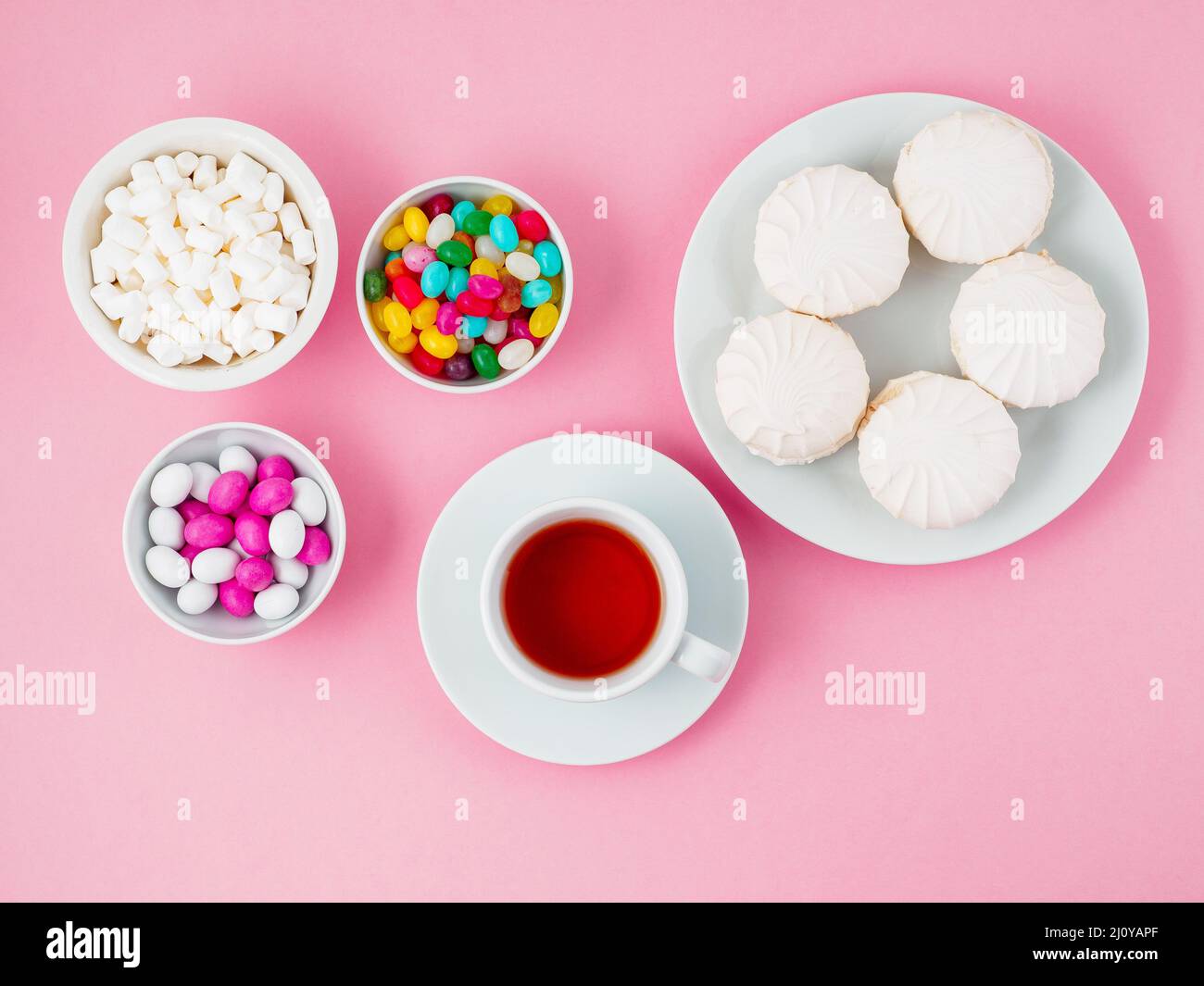 Tasse Tee, Teller mit verschiedenen Süßigkeiten-Marshallows, Lutscher, Süßigkeiten. Kohlenhydrate, Glukose. Rosafarbener Hintergrund, flach liegend. Stockfoto