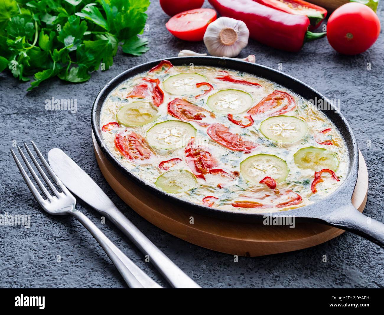 Pfanne mit traditionellen italienischen Frittata auf dunkelgrauem Steintisch, Gabeln, Petersilie, Pfeffer, Tomaten. Rühreier Stockfoto