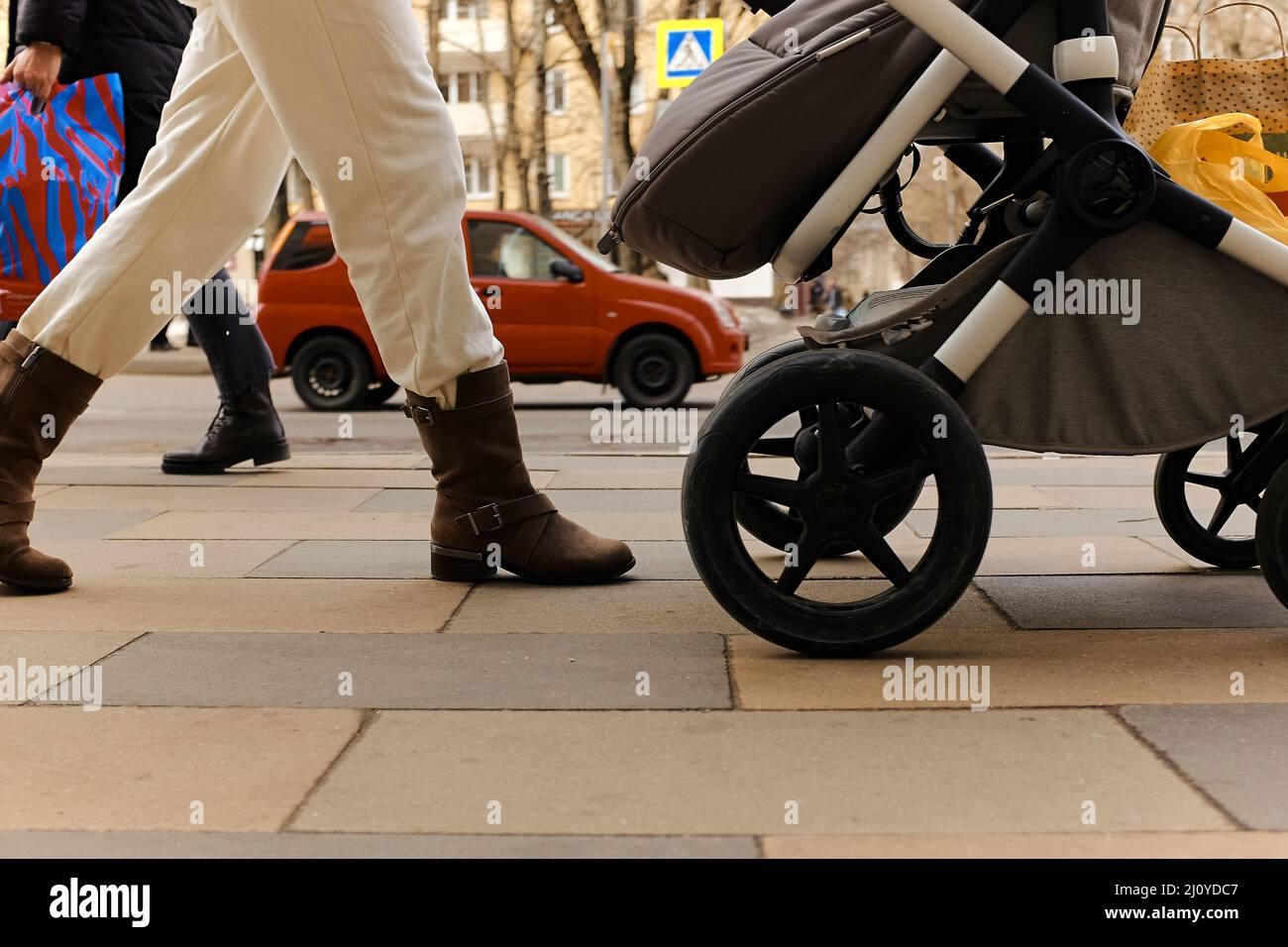Stilvolle Frau geht mit Kinderwagen, geht mit Einkäufen aus dem Geschäft, rotes Auto fährt vorbei, viel Verkehr auf dem Bürgersteig Stockfoto
