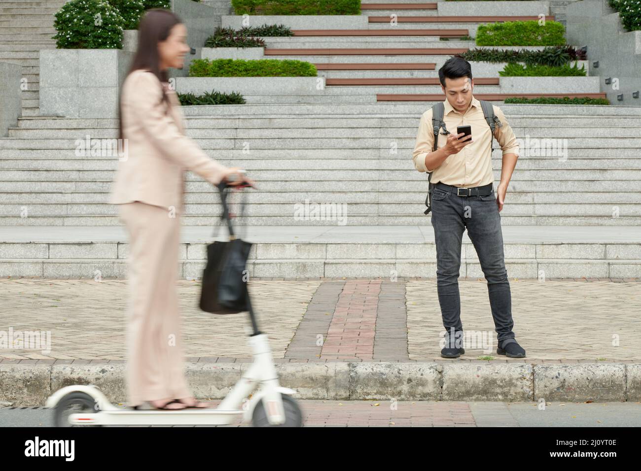 Ernsthafter junger Mann, der auf dem Bürgersteig steht und SMS auf dem Smartphone liest, wenn eine Frau auf dem Roller vorbeifährt Stockfoto
