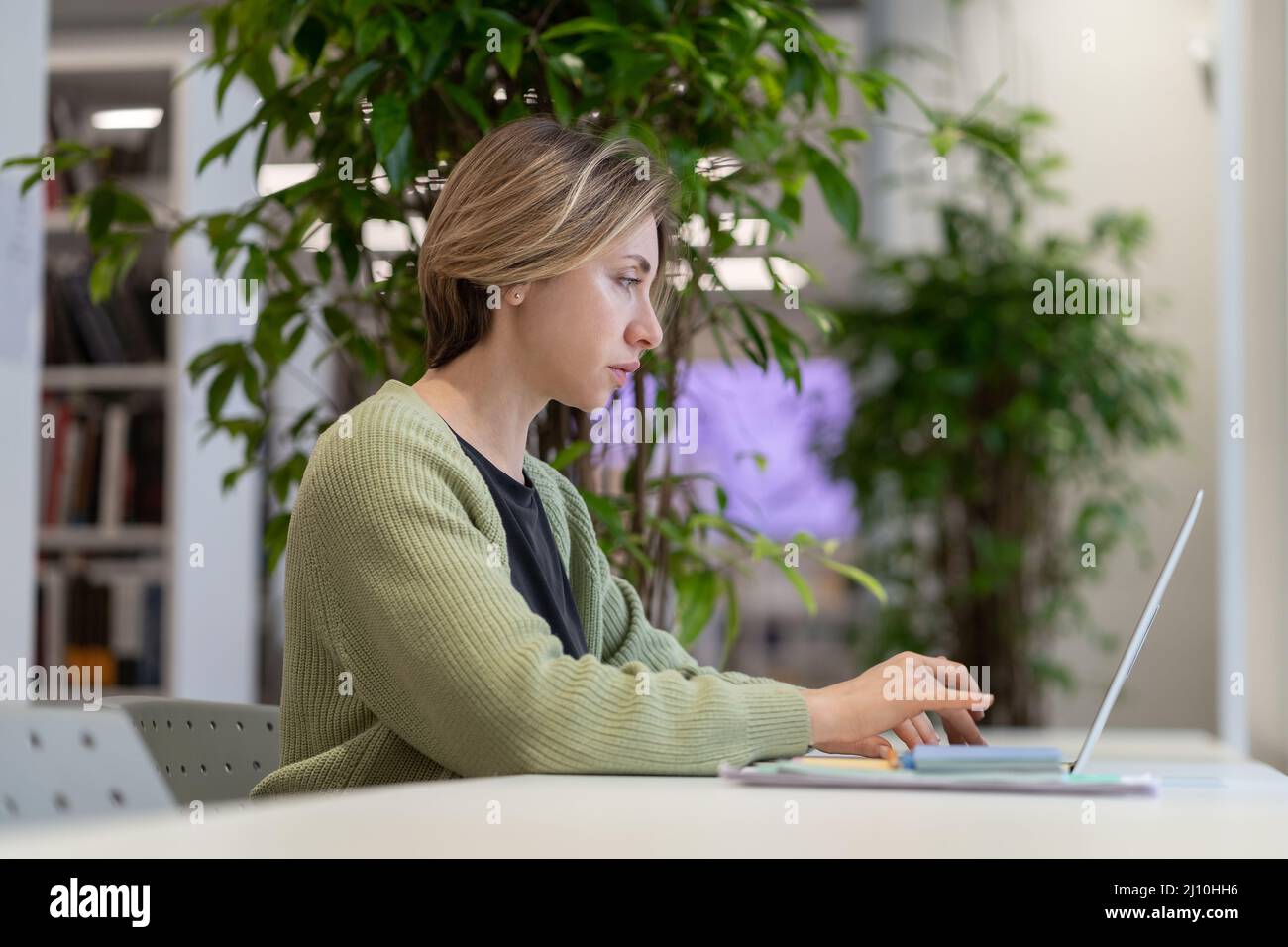 Nachdenkliche Universitätslehrerin, die in einer gemütlichen leeren Bibliothek mit grünen Zimmerpflanzen am Laptop arbeitet Stockfoto