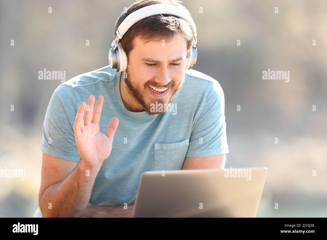 Fröhlicher Mann mit Kopfhörern, der während eines Videoanrufs auf einem Laptop im Freien mit der Hand begrüßt Stockfoto