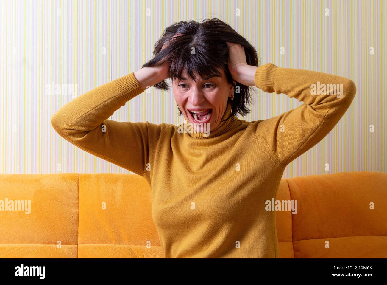 Verärgert schreiende Frau mittleren Alters, die unter Stress leidet und ihren Kopf mit den Händen auf einem orangefarbenen Sofa hält. Krise des mittleren Alters. Konzept der psychischen Gesundheit. Stockfoto