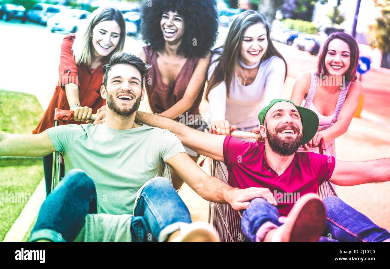 Junge multirassische Freunde, die zusammen mit dem Einkaufswagen Spaß haben - Millennials, die sich Zeit mit Trolleys auf dem Stadtparkplatz eines Einkaufszentrums teilen - Anm. Stockfoto
